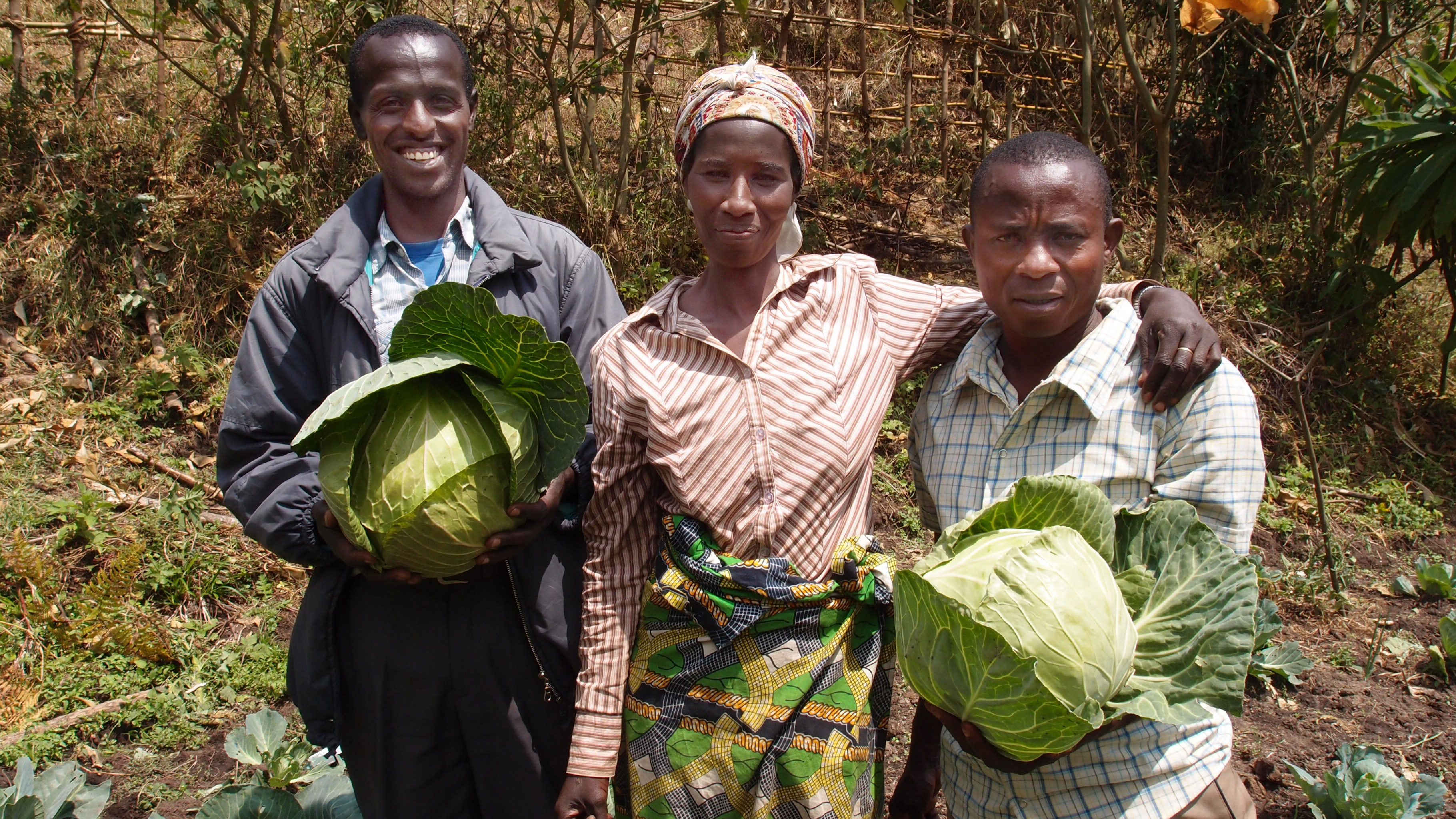 Die 40-Jährige Perusi Nyarukund (m) hat Gemüsekulturen erhalten und begleitet von dem landwirtschaftlichen Berater Aimable (li) einen Gemüsegarten angelegt. Der Projekt-Agronom von UGEAFI, Ilelo A-umba Leli (re), hat den Zaun für den Garten gebaut.