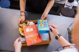 Eine kleine Kiste mit Sachspenden, u.a. einem Kochbuch und einem Spiel, wird im Oxfam Shop abgegeben.