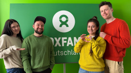 Laura, Florian, Carmen und Charlie stehen vor einem Oxfam-Logo und tragen verschieden farbige Pullover von Organicforce.