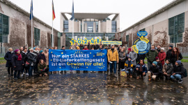 Aktivist*innen der "Initiative Lieferkettengesetzt" mit einem Banner „Nur ein starkes EU-Lieferkettengesetz ist ein Gewinn für alle!“ vor dem Bundeskanzleramt. In der hinteren Reihe halten einige Aktivist*innen die Anzahl der eingereichten Unterschriften (90.248) hoch. Rechts ist ein Glücksrad zu sehen, das den „Gewinn für alle“ als Hauptgewinn anzeigt.