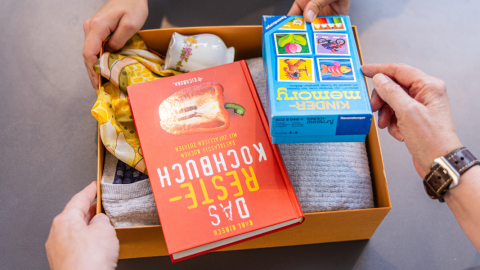 Eine kleine Kiste mit Sachspenden, u.a. einem Kochbuch und einem Spiel, wird im Oxfam Shop abgegeben.