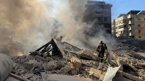 Die Trümmer eines Hauses zwischen anderen Gebäuden. Es steigt Rauch auf. Zwei Menschen bewegen sich über die Trümmer.