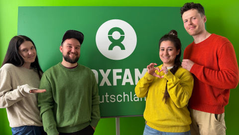 Laura, Florian, Carmen und Charlie stehen vor einem Oxfam-Logo und tragen verschieden farbige Pullover von Organicforce.
