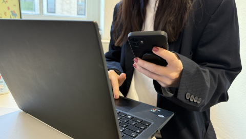 Pressekontakt Symbolbild: Eine Frau steht vor einem Laptop und hält ein Handy in der Hand.